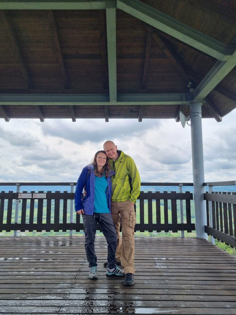 Para turystów uśmiechnięta pozuje na drewnianym tarasie widokowym zadaszonej wieży na Szpilówce. W tle panorama zielonych wzgórz i pochmurne niebo.