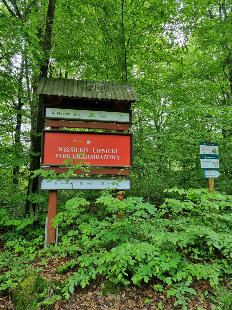 Drewniana tablica informacyjna przy wejściu do Wiśnicko-Lipnickiego Parku Krajobrazowego, otoczona gęstym zielonym lasem.