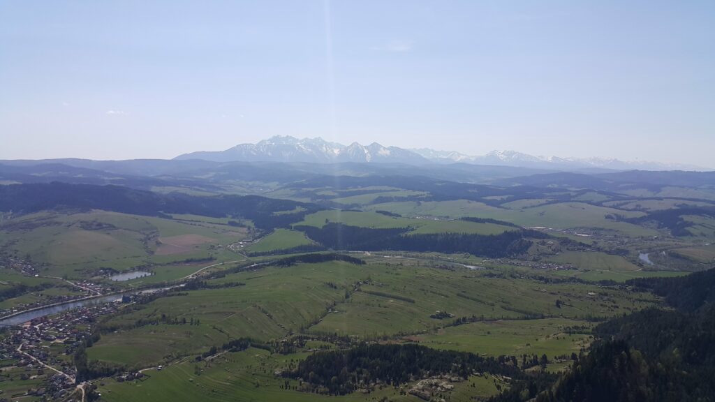 Panorama widziana ze szczytu Trzy Korony w Pieninach, z rozległym krajobrazem dolin i ośnieżonymi Tatrami w tle.