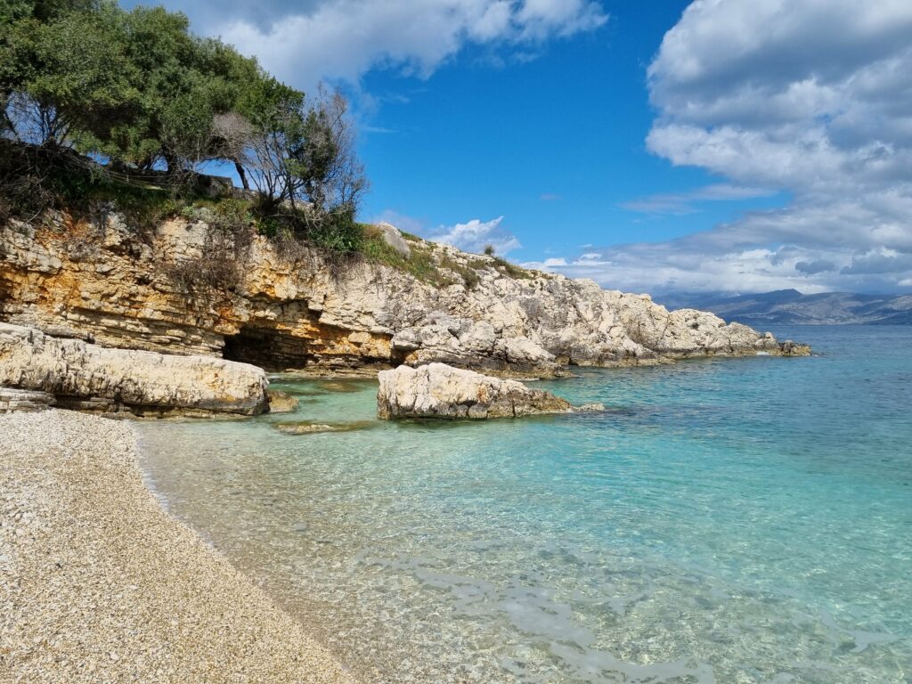 Kamienista plaża z turkusową wodą i jaskinią w skale na Korfu