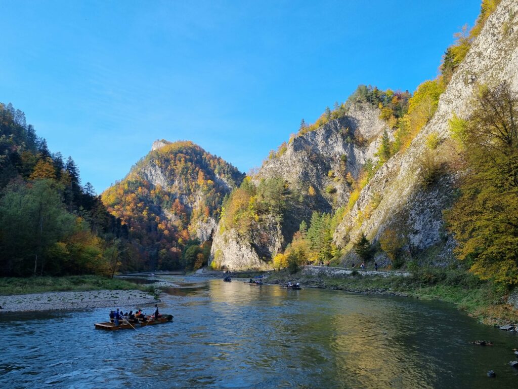 Jesienna panorama przełomu Dunajca z tratwą flisacką na rzece i ścieżką rowerową biegnącą wzdłuż skalistego brzegu.