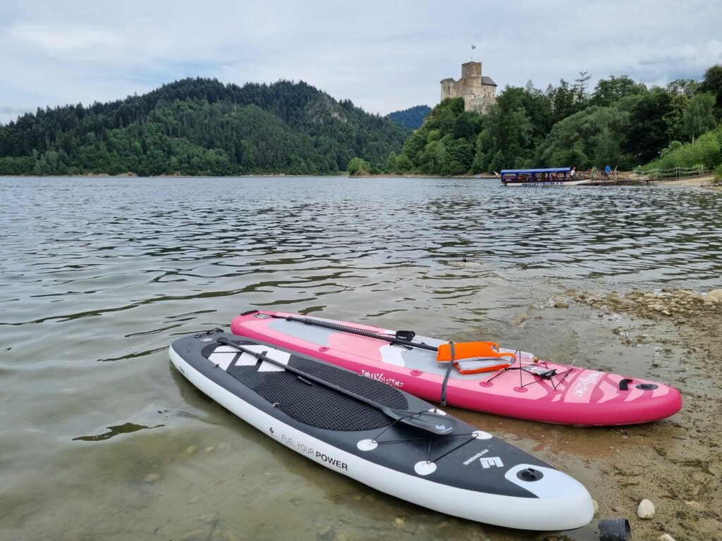 Dwie deski SUP, różowa i czarna, leżące na piaszczystym brzegu Jeziora Czorsztyńskiego, z Zamkiem w Niedzicy widocznym w oddali na zalesionym wzgórzu.