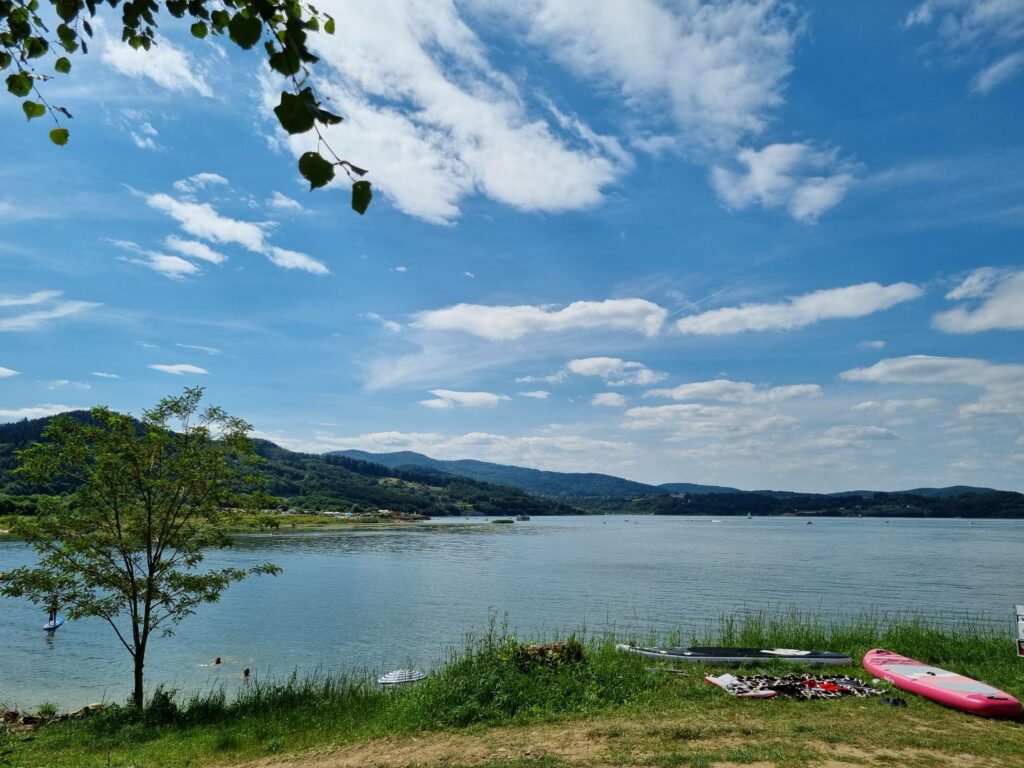 Dwie deski SUP leżące na trawiastym brzegu Jeziora Mucharskiego, z rozległą taflą wody i zalesionymi wzgórzami w tle pod niebieskim niebem z chmurami.