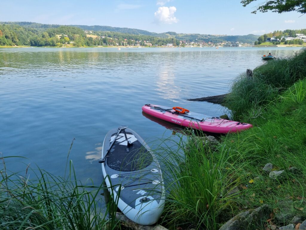 Dwie deski SUP, różowa i czarno-biała, częściowo w wodzie przy trawiastym brzegu Jeziora Rożnowskiego, z zabudowaniami i pagórkami w tle pod jasnym niebem.