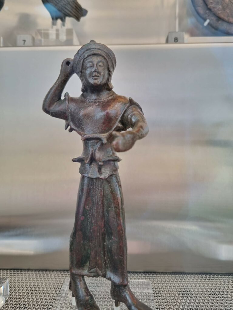 Mała, starożytna figurka z brązu, przedstawiająca postać ludzką, eksponowana w muzeum na szklanej półce.