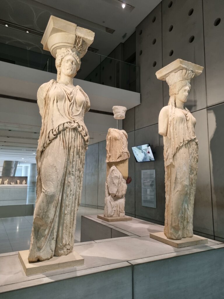 Oryginalne rzeźby kariatyd z Erechtejonu, eksponowane w nowoczesnym Muzeum Akropolu w Atenach, prezentowane w przestronnej sali z naturalnym światłem.