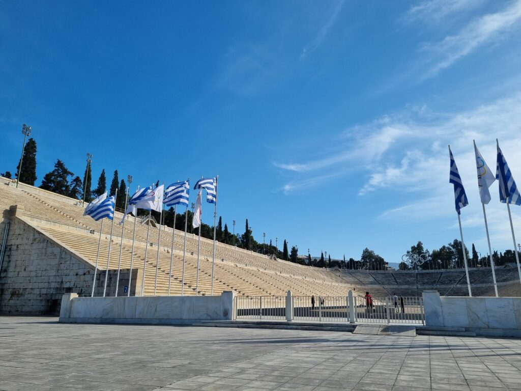 Panathenaic Stadium w Atenach, pierwszy nowożytny stadion olimpijski, z marmurowymi trybunami i powiewającymi flagami Grecji oraz flagami olimpijskimi pod błękitnym niebem.
