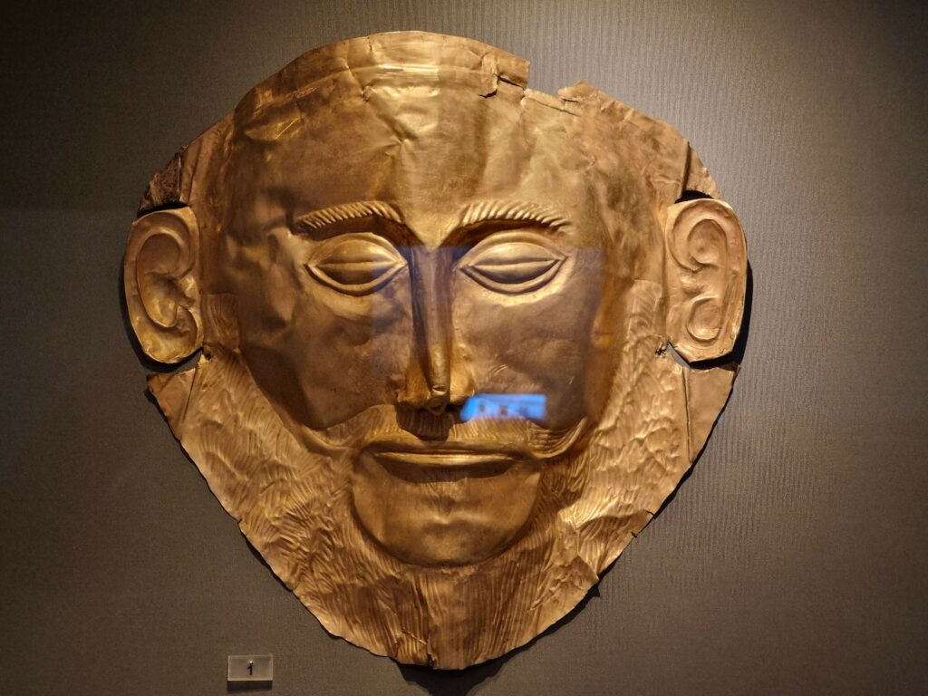Złota Maska Agamemnona, starożytny artefakt pogrzebowy wykonany z litego złota, z detalami twarzy i zarostu, eksponowana w muzeum na ciemnym tle.