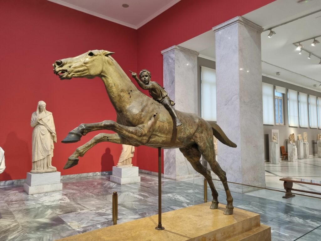 Duża, brązowa rzeźba konia z młodym jeźdźcem, eksponowana w Archeologicznym Muzeum Narodowym w Atenach, w przestronnej sali z marmurowymi posągami w tle.