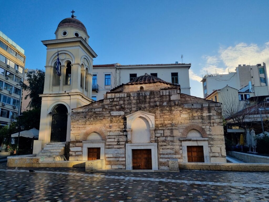 Starożytny kościół Panagia Kapnikarea w Atenach z kamienną fasadą i dzwonnicą, położony na mokrym placu miejskim pod błękitnym niebem.