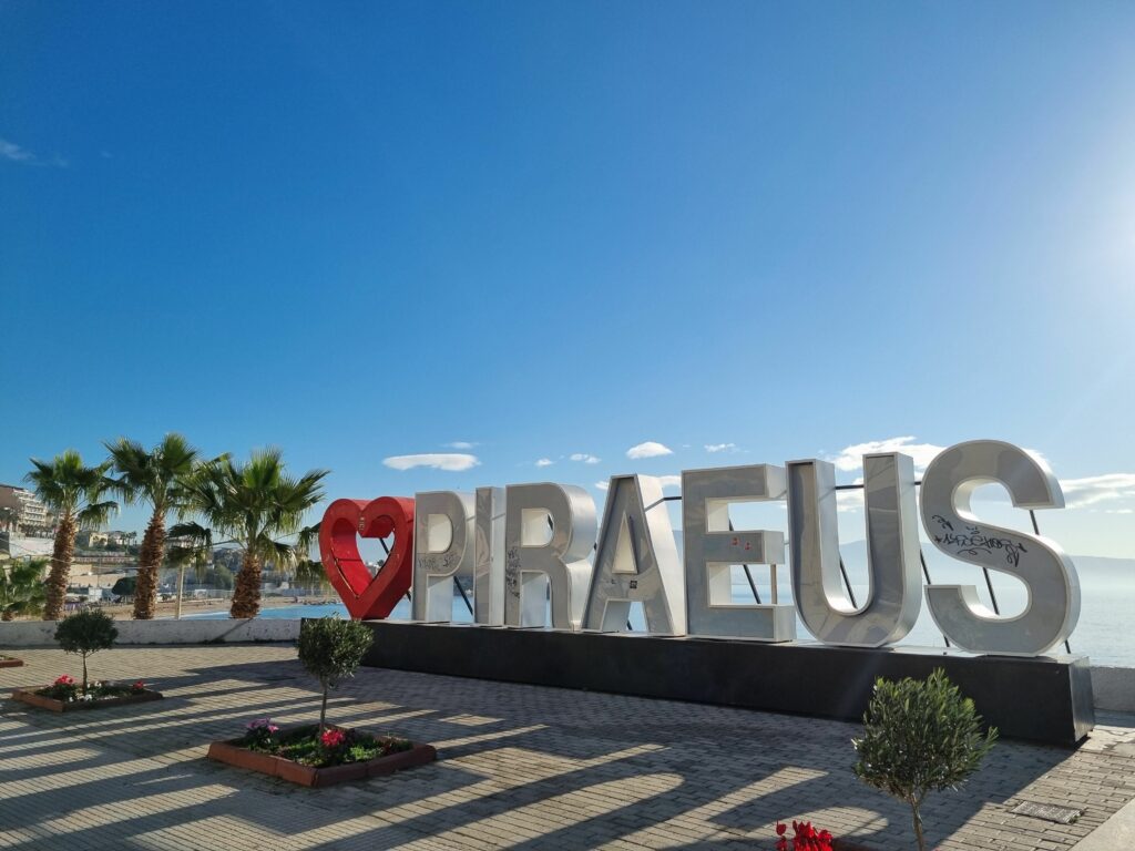 Duży, biały napis "I LOVE PIRAEUS" (z sercem zamiast "LOVE") na promenadzie w Pireusie, z palmami, morzem i budynkami w tle pod błękitnym niebem.