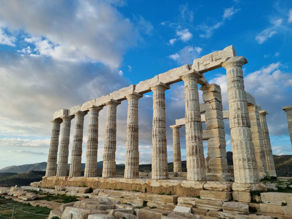 Ruiny starożytnej Świątyni Posejdona w Sounion, z imponującymi kolumnami stojącymi na cyplu, na tle błękitnego nieba z chmurami i morza w oddali.