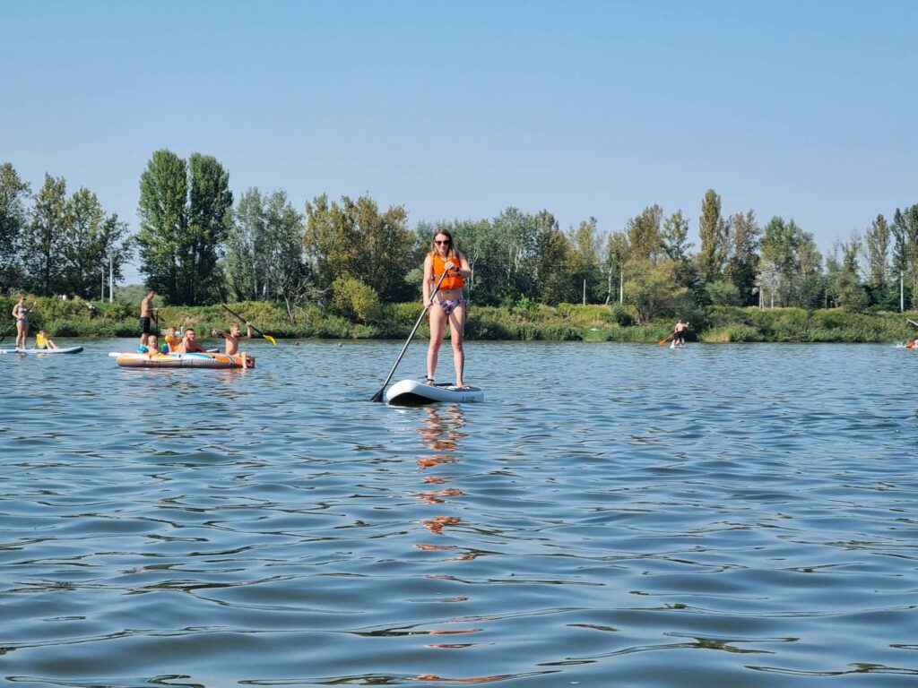 Kobieta w pomarańczowej kamizelce i stroju kąpielowym stoi na desce SUP na Zalewie Przylasek Rusiecki, a w tle widać innych ludzi na SUPach i kajakach oraz zalesiony brzeg.