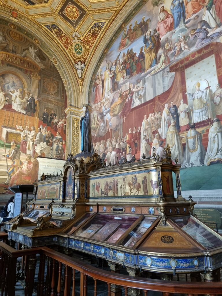 Bogato zdobione wnętrze Muzeów Watykańskich z monumentalnymi freskami ściennymi przedstawiającymi sceny religijne oraz dużą, ozdobną witryną ekspozycyjną z drobnymi przedmiotami.