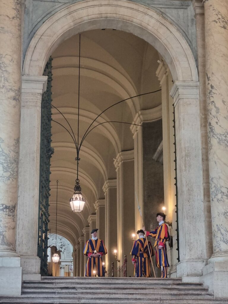 Członkowie Gwardii Szwajcarskiej w tradycyjnych, kolorowych uniformach, stojący na straży w łukowatym korytarzu Watykanu, z rzeźbionymi kolumnami i wiszącymi lampami.