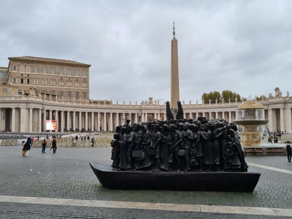 Plac św. Piotra w Watykanie z brązową rzeźbą "Angels Unawares" przedstawiającą grupę migrantów na łodzi, z obeliskiem i kolumnadą w tle pod pochmurnym niebem.