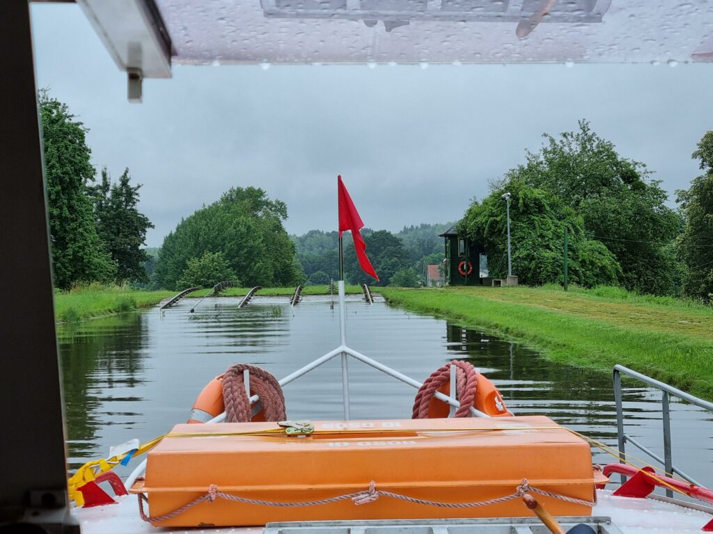 Perspektywa z przodu statku na Kanał Elbląski, z czerwoną flagą na dziobie.