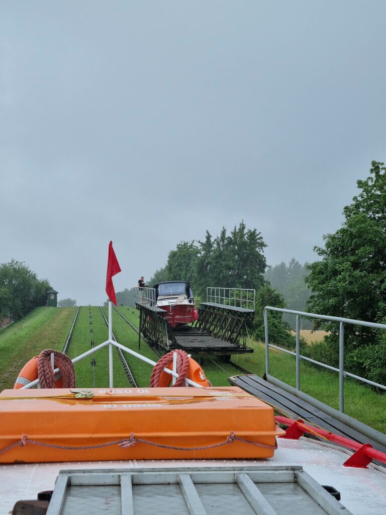 Widok z dziobu statku na pochylnię Kanału Elbląskiego z inną łodzią na wózku w oddali, pokonującą wzniesienie.