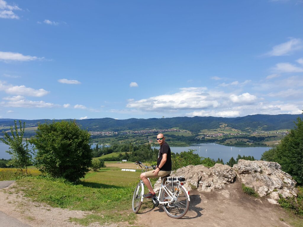 Rowerzysta na szlaku Velo Czorsztyn, z widokiem na Jezioro Czorsztyńskie i góry.