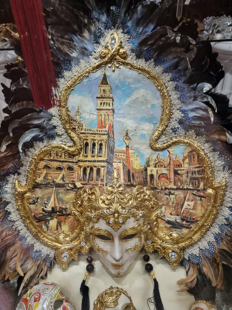 Bogato zdobiona wenecka maska karnawałowa z piórami i inkrustacjami, z obrazem przedstawiającym Plac Świętego Marka i dzwonnicę w tle.