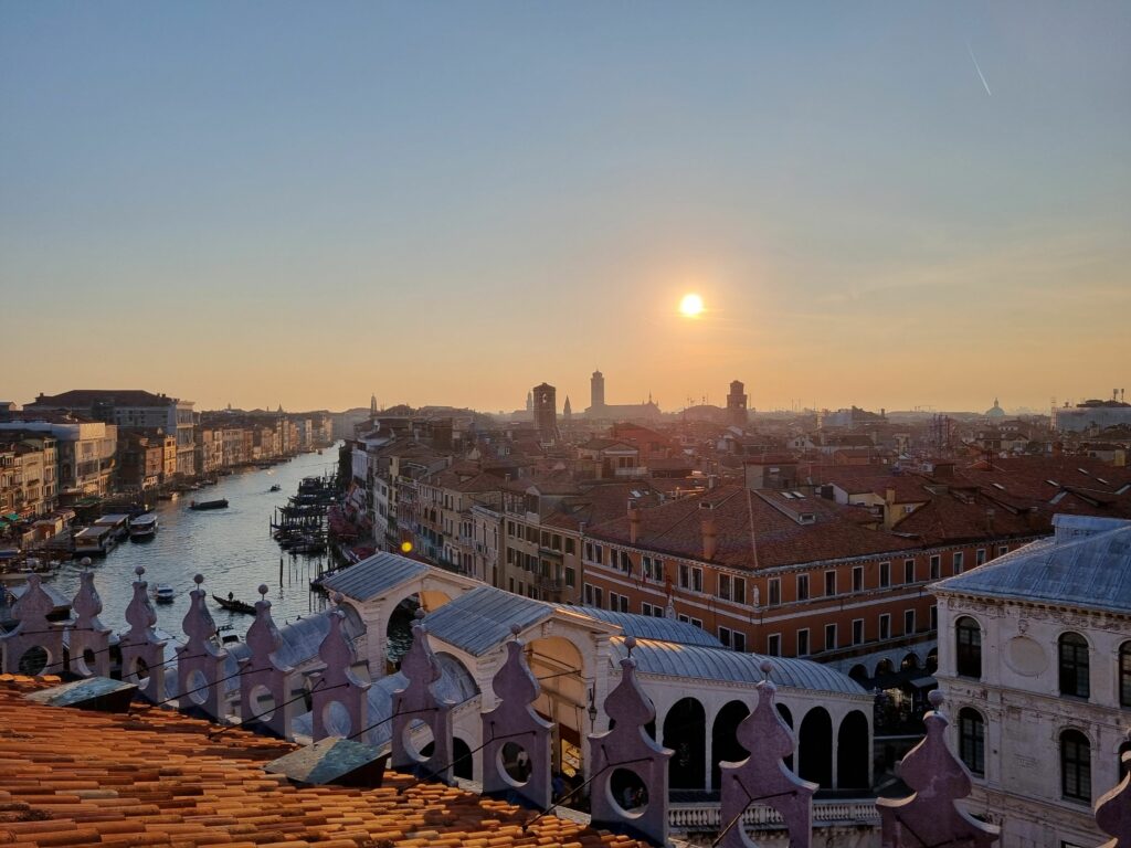 Panoramiczny widok na Wenecję o zachodzie słońca, z Canal Grande wijącym się między dachami zabytkowych budynków, oświetlonym ciepłym, pomarańczowym blaskiem słońca.