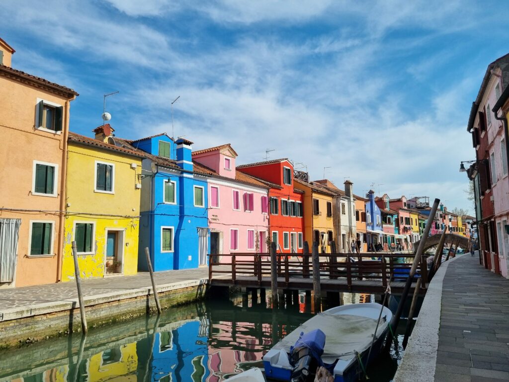 Malowniczy kanał na Burano otoczony jaskrawo kolorowymi domami, z drewnianym mostkiem łączącym brzegi i łodziami zacumowanymi w wodzie, pod dynamicznym, częściowo zachmurzonym niebem.