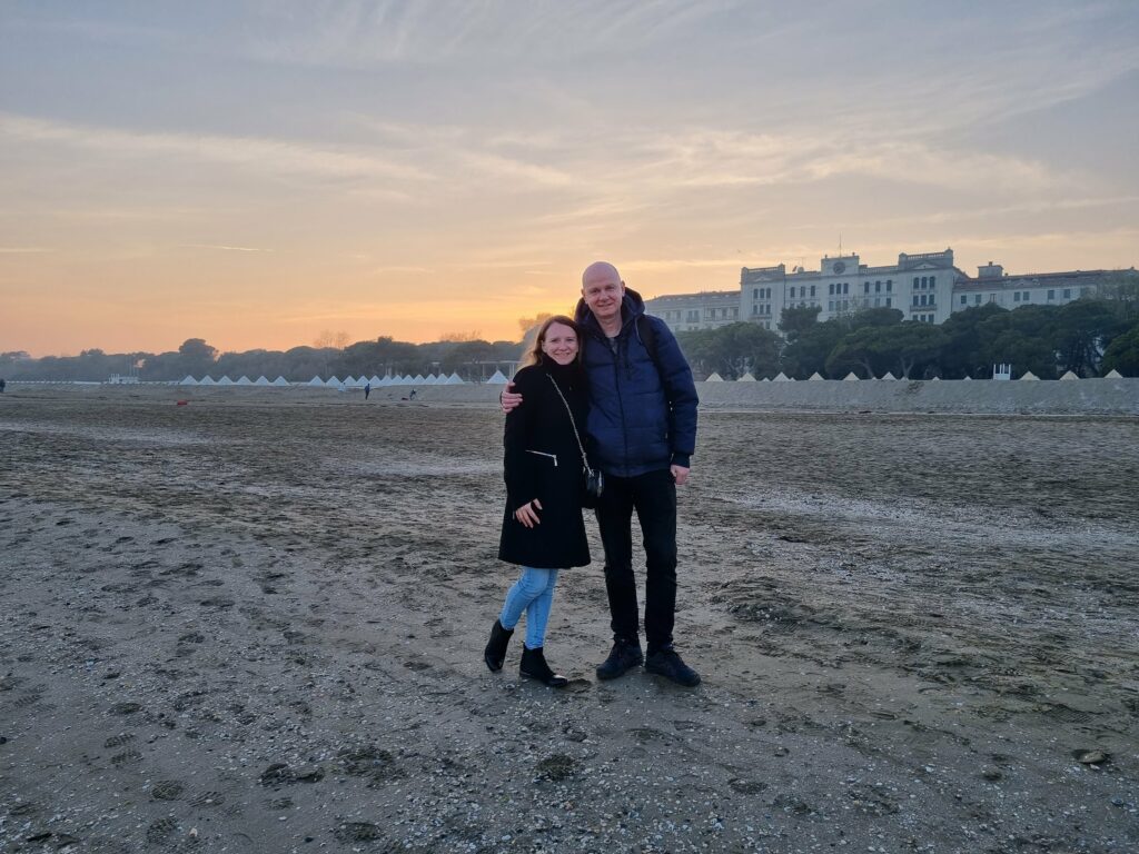 Para stojąca na szerokiej, piaszczystej plaży na wyspie Lido o zachodzie słońca, z rozproszonym światłem i dużym budynkiem w tle.
