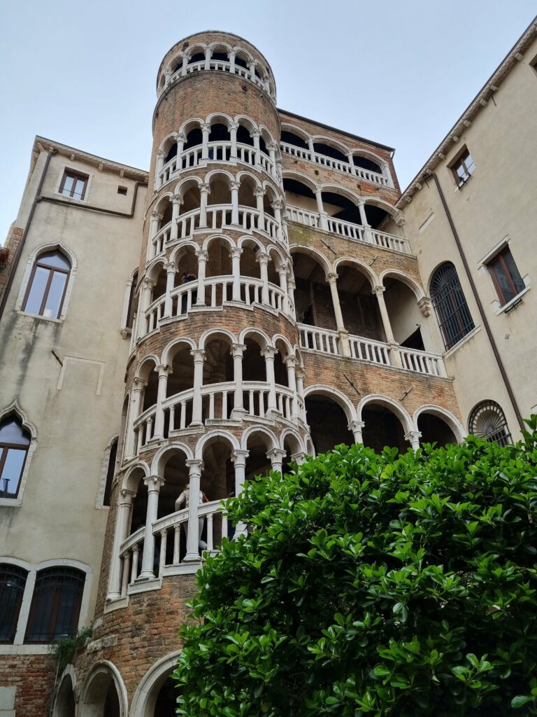 Wysoka, cylindryczna wieża ze spiralnymi arkadowymi schodami Palazzo Contarini del Bovolo w Wenecji, otoczona zabytkowymi budynkami i zielonymi krzewami.