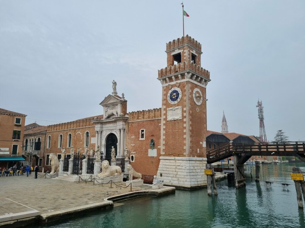 Historyczne wejście do Arsenale di Venezia z monumentalną bramą, wieżą zegarową i rzeźbami lwów, otoczone wodą kanału i pobliskim drewnianym mostem.