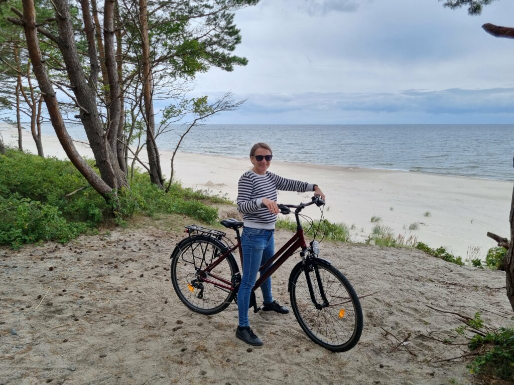 Uśmiechnięta kobieta na rowerze, na tle morza i plaży na Mierzei Wiślanej.