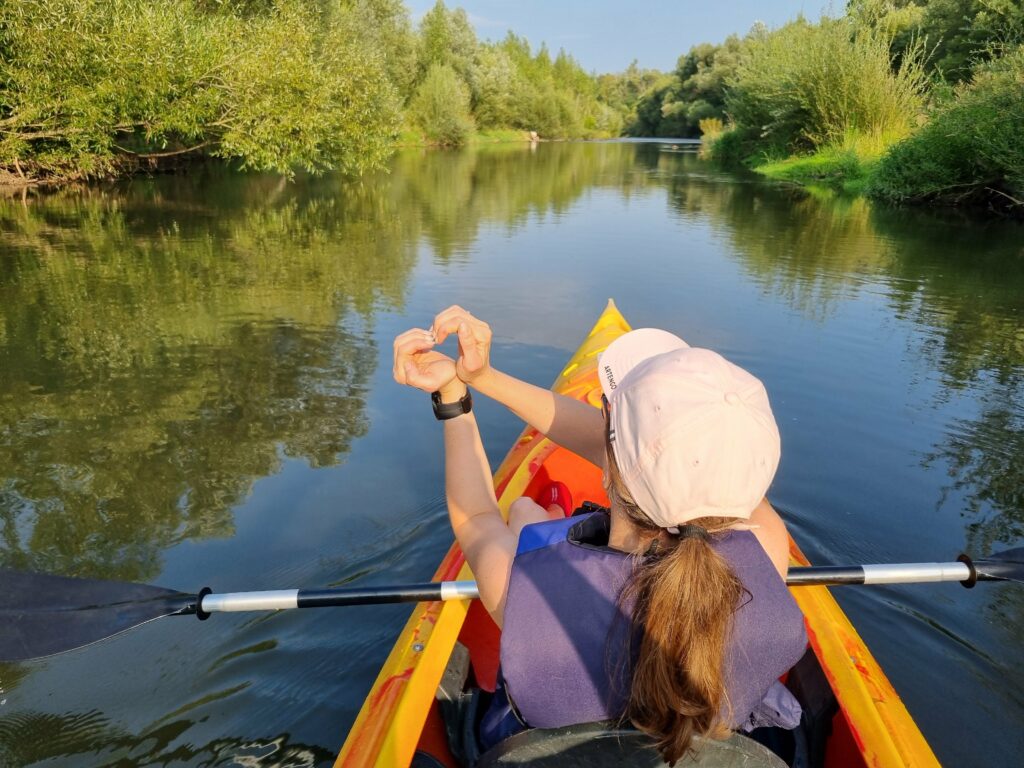 Kobieta w kajaku, widziana od tyłu, układa dłonie w kształt serca. W tle widać rzekę Raba i zielone drzewa, a słońce odbija się od wody.