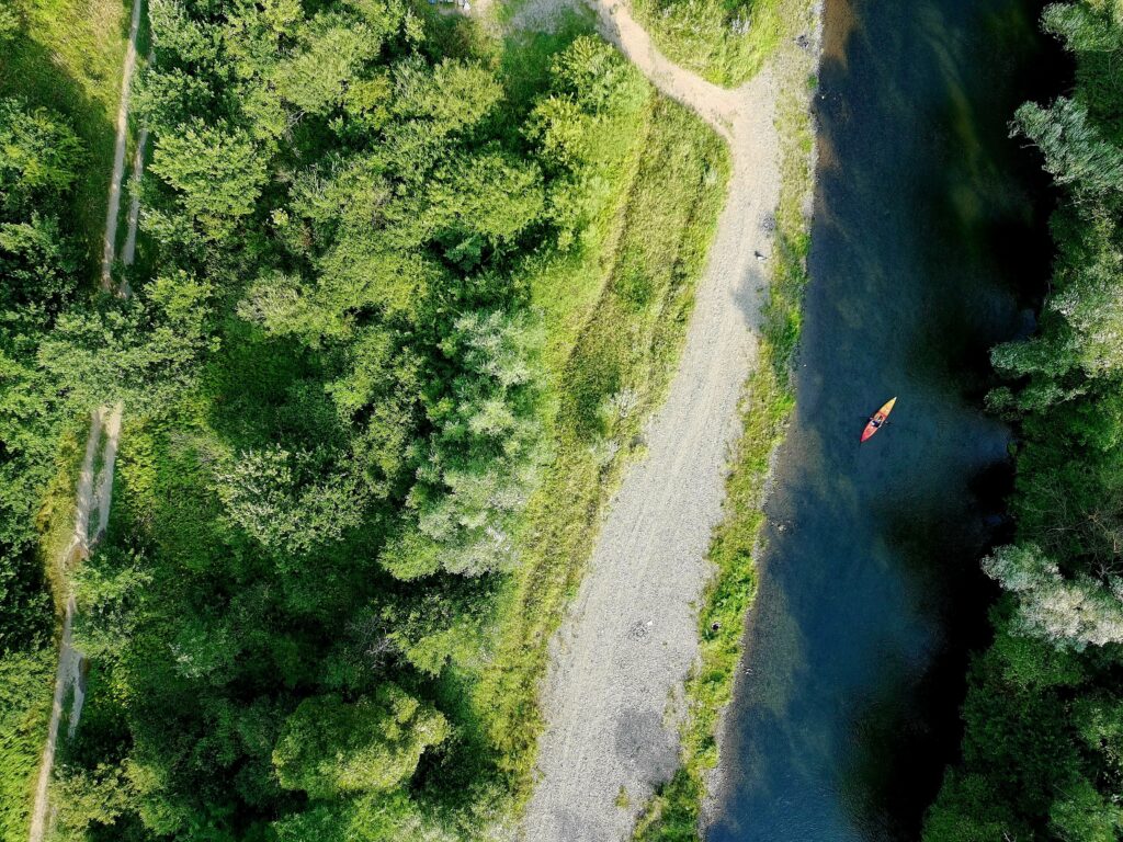 Ujęcie z drona na jeden kajak płynący rzeką Raba, otoczoną gęstą roślinnością i ścieżkami na brzegu.