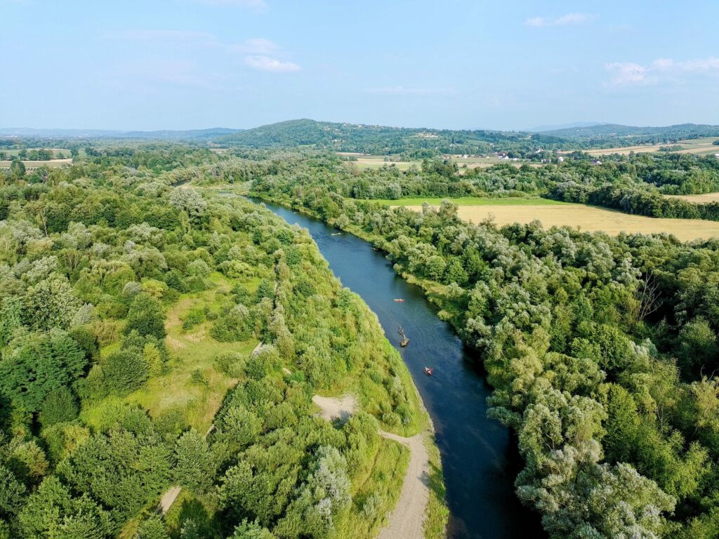 Szeroki widok z drona na rzekę Raba, wijącą się przez dolinę otoczoną lasami, polami i wzgórzami. Na rzece widoczne są małe kajaki.