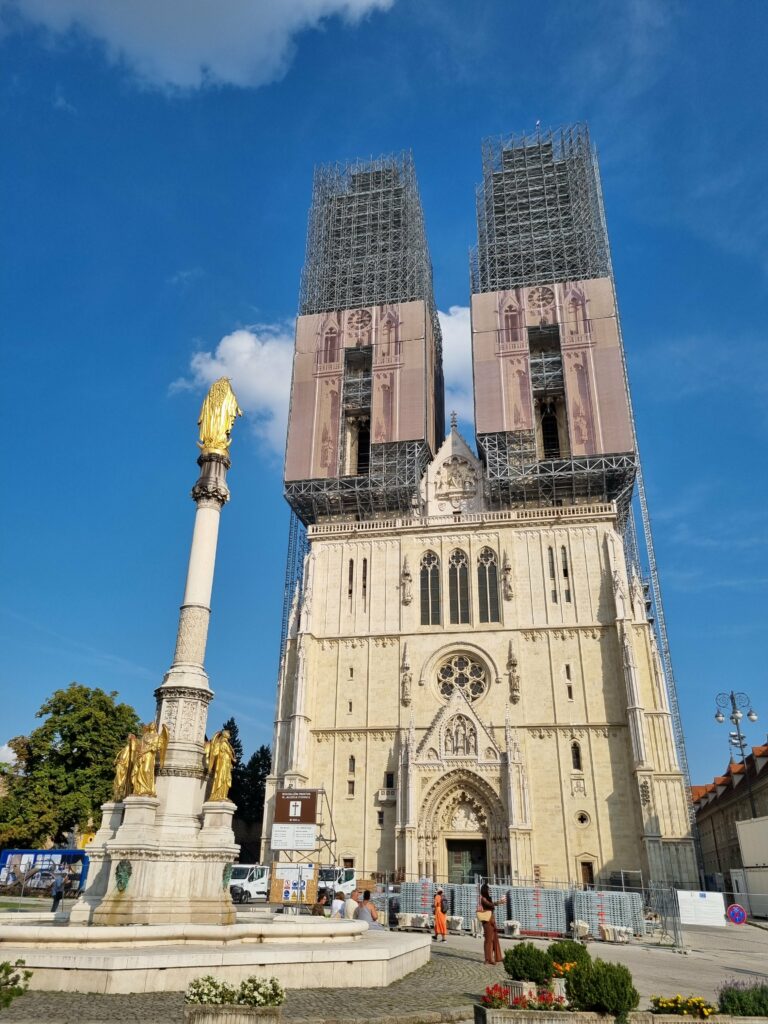 Katedra w Zagrzebiu z wieżami w remoncie, widok od frontu. Przed nią znajduje się kolumna z figurą Najświętszej Maryi Panny na tle błękitnego nieba.