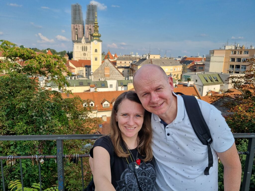 Uśmiechnięta para turystów robiąca selfie na punkcie widokowym w Górnym Mieście. W tle panorama Zagrzebia z widokiem na wieże Katedry.