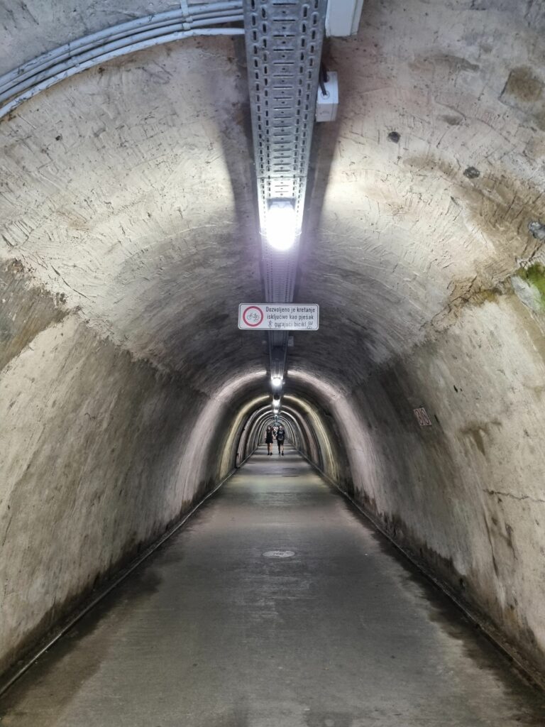 Wnętrze tunelu Grič w Zagrzebiu. Długi, betonowy korytarz o łukowatym sklepieniu, oświetlony lampami, w oddali widać sylwetki ludzi.