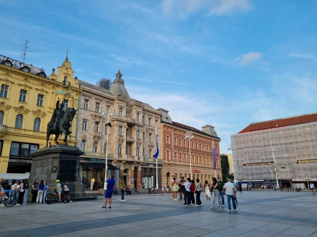 Szeroki widok na Plac Bana Jelačicia w Zagrzebiu w późne popołudnie. Na placu widać pomnik konny, otaczające go zabytkowe kamienice i spacerujących ludzi.
