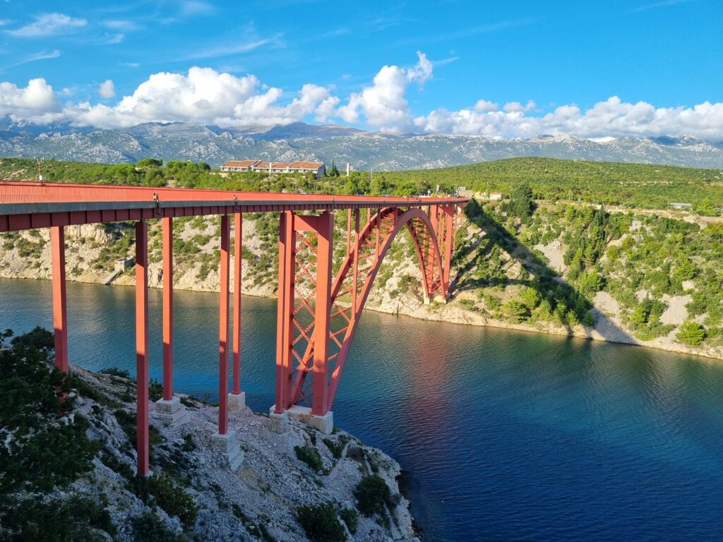 Czerwony most Maslenica nad błękitną wodą w Chorwacji, w tle góry Velebit i błękitne niebo.