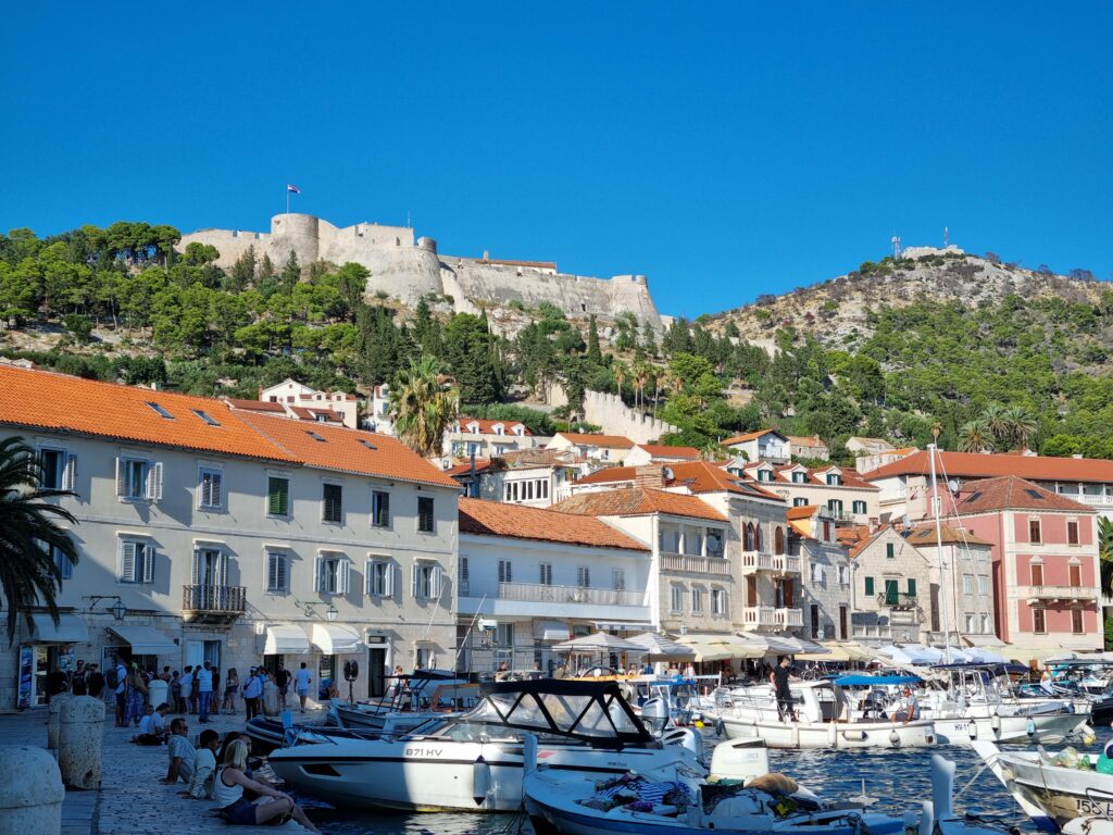 Tętniący życiem port w mieście Hvar. Liczne jachty i łodzie zacumowane przy promenadzie, nad którą góruje historyczna twierdza Fortica (Španjola) na zielonym wzgórzu.