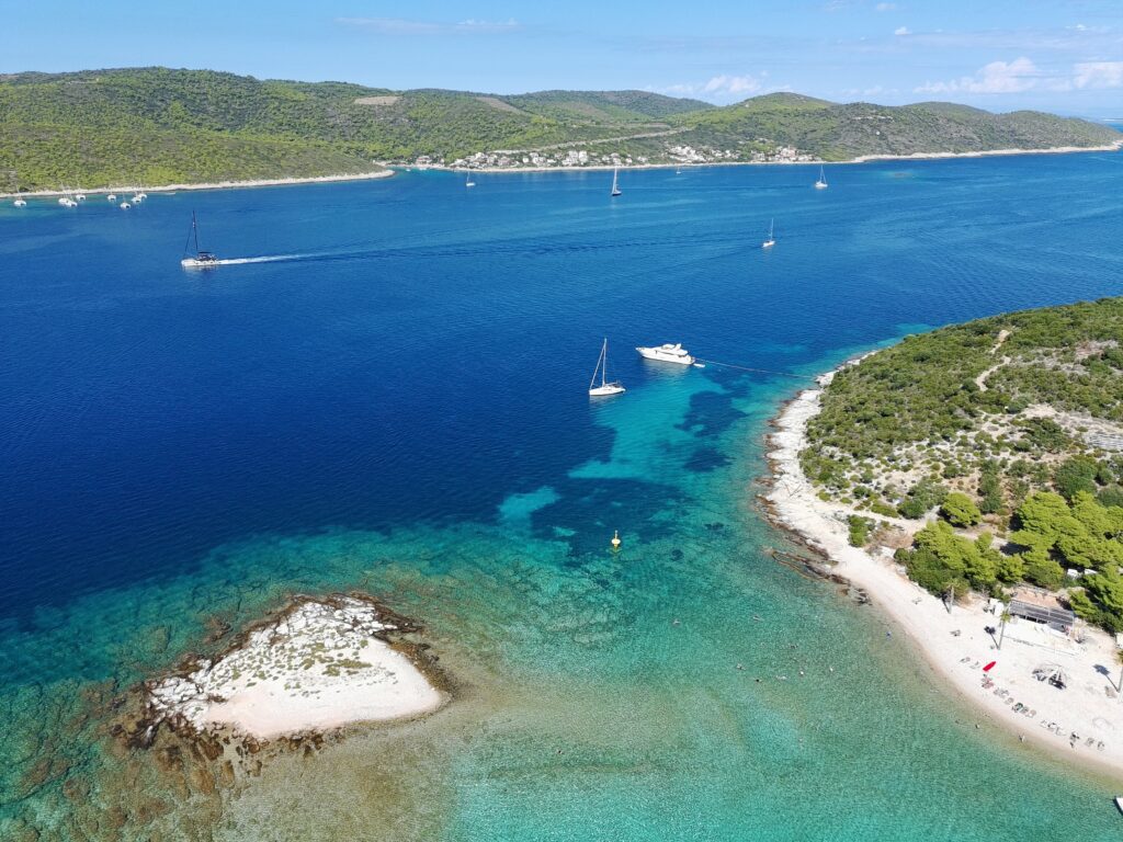 Szeroka panorama z drona na wyspę Budikovac i otaczającą ją Błękitną Lagunę. Widać piaszczystą plażę z leżakami, a na błękitnym morzu płyną jachty i żaglówki na tle zielonych wysp Dalmacji.
