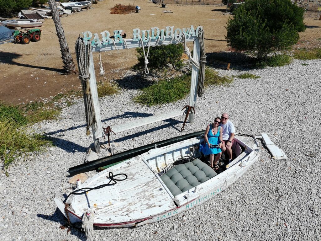 Para turystów siedząca w starej, białej łodzi ustawionej na kamienistej plaży wyspy Budikovac. Za nimi znajduje się drewniana rama z napisem "PARK BUDIKOVAC", popularne miejsce do robienia zdjęć.