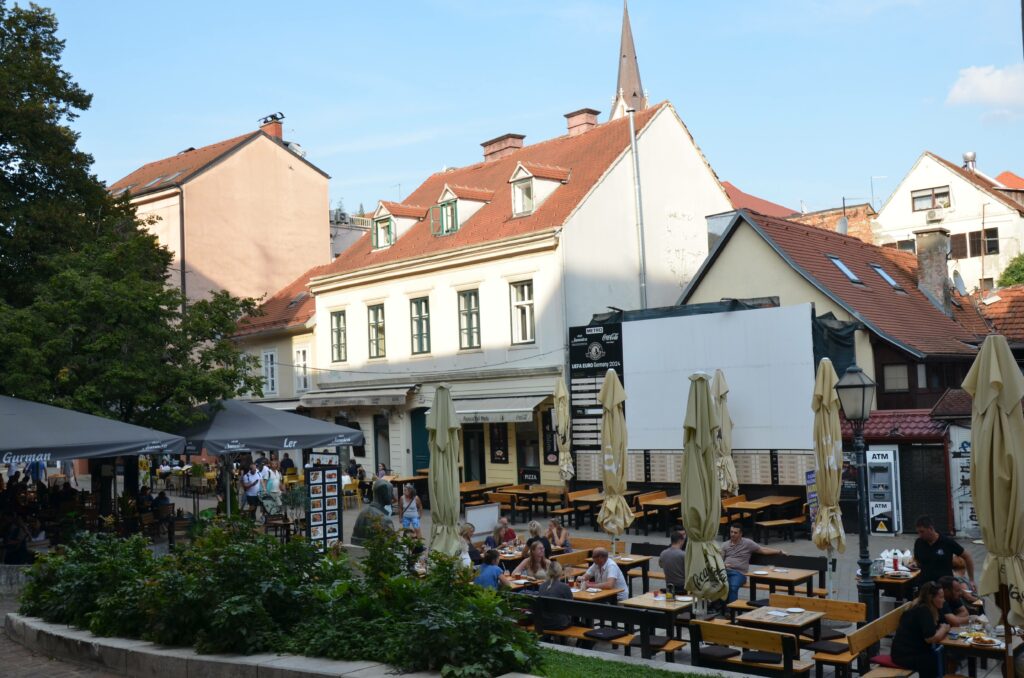 Ogródki kawiarniane na jednym z placów w Zagrzebiu, wypełnione ludźmi siedzącymi przy stolikach. W tle widać historyczne kamienice i wieżę kościoła.