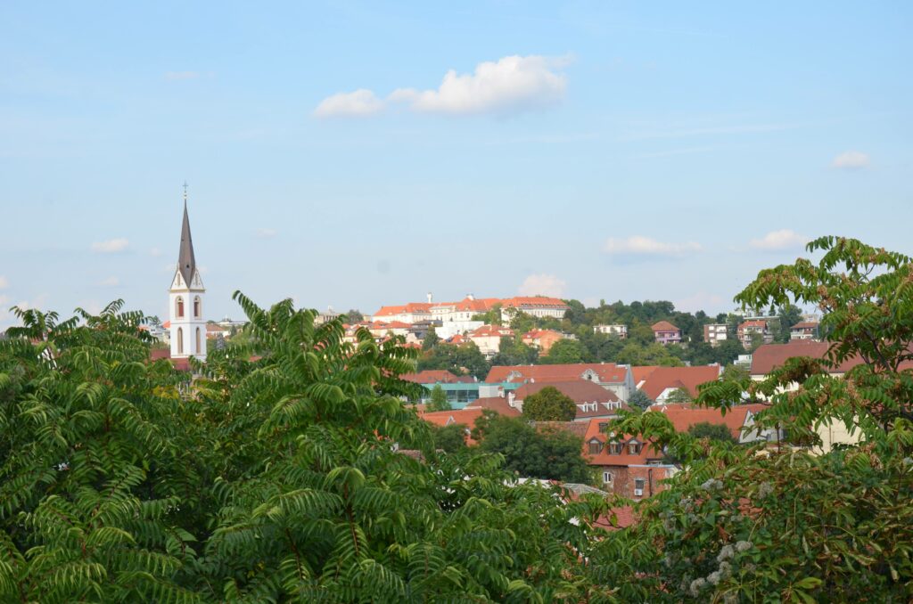 Panorama Zagrzebia widziana z Górnego Miasta. Na pierwszym planie zielone drzewa, dalej czerwone dachy kamienic i wieża kościoła na tle błękitnego nieba.
