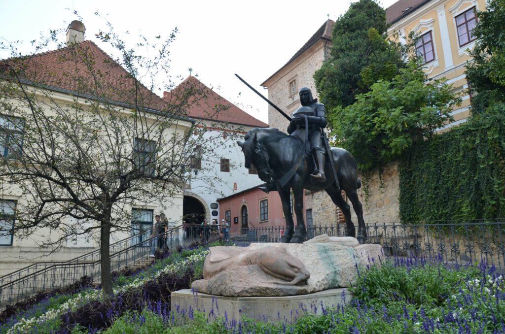 Brązowy pomnik konny św. Jerzego walczącego ze smokiem, znajdujący się w Górnym Mieście w Zagrzebiu, w otoczeniu kwiatów i historycznych kamienic.
