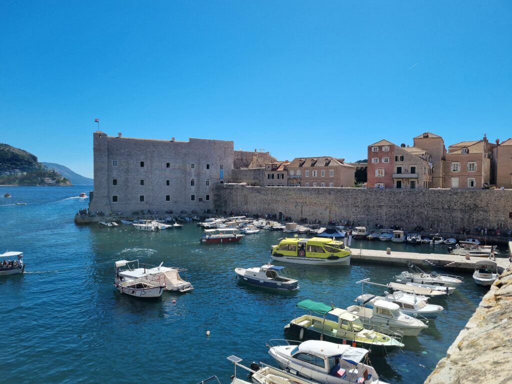 Stary Port w Dubrowniku wypełniony łódkami, widoczny Fort św. Jana i mury obronne.