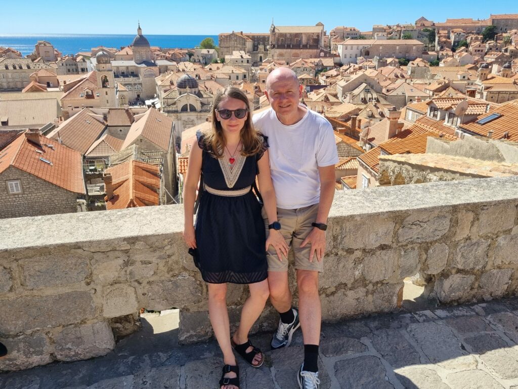Para turystów na murach miejskich Dubrownika, w tle panorama czerwonych dachów Starego Miasta i katedra.