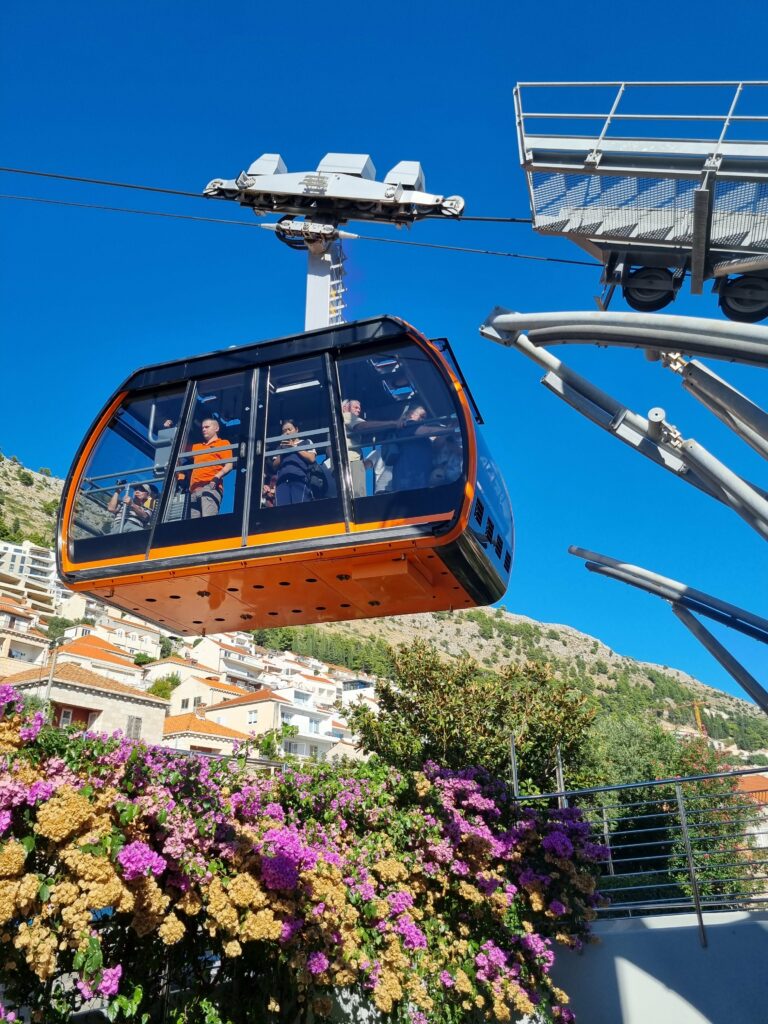 Pomarańczowa gondola kolejki linowej wjeżdżająca na wzgórze Srđ nad Dubrownikiem, na pierwszym planie kwitnące bugenwille.