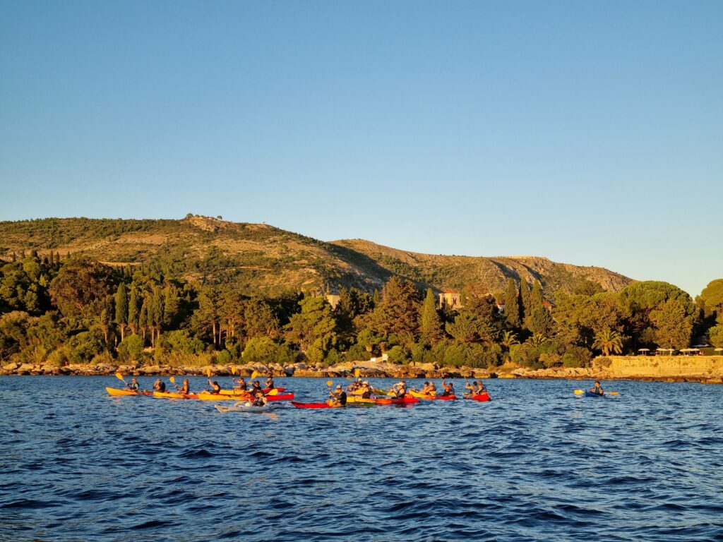 Grupa turystów w kajakach morskich wiosłująca wzdłuż wybrzeża Dubrownika o zachodzie słońca.