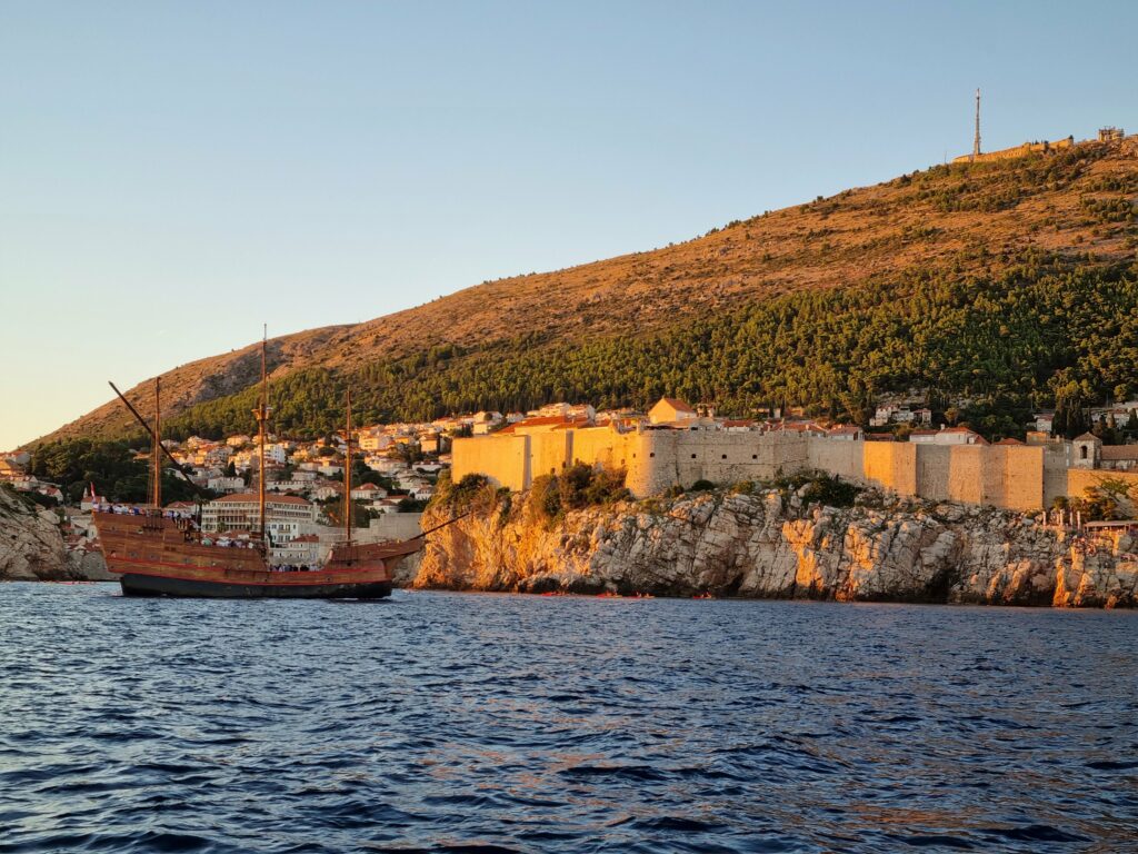 Drewniany statek stylizowany na dawną karakę płynie na tle oświetlonych zachodzącym słońcem murów miejskich Dubrownika.