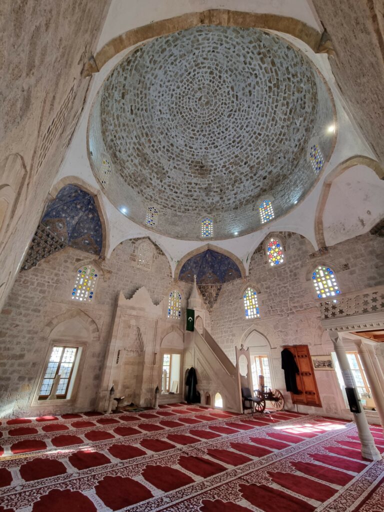 Wnętrze meczetu Hadži-Alija w Počitelj z kamienną kopułą, witrażami i tradycyjnym czerwonym dywanem modlitewnym.
